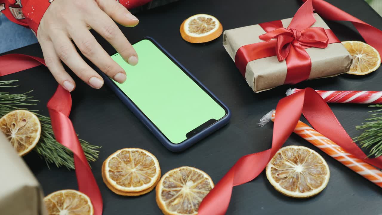 la mujer está buscando regalos de navidad en línea en su teléfono inteligente con pantalla verde, llave de croma. compras navideñas. la mujer está sentada en casa en una habitación decorada en navidad y mirando las redes sociales, desplazando las noticias