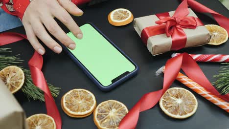 여성은 녹색 화면, 크로마 키, 휴가 쇼핑을 가진 스마트폰에서 온라인에서 xmas 선물을 검색하고 있습니다. 여성은 크리스마스에 장식된 방에서 집에 앉아 소셜 미디어를보고 뉴스를 스크롤하고 있습니다.