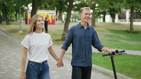 The-guy-and-the-girl-are-walking-in-the-park,-talking.-The-guy-is-holding-a-scooter-in-his-hand.-Happy-walk.-romantic-date