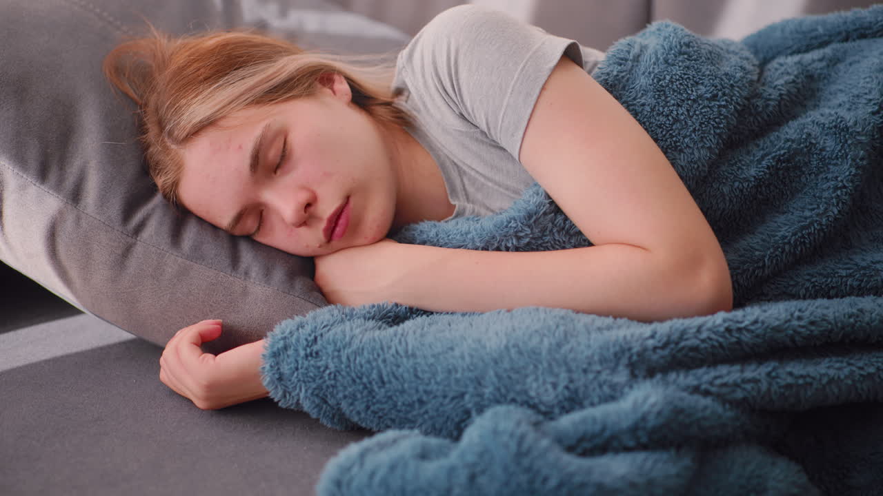 Close up of young woman sleeping peacefully on couch with head resting on grey cushion, hand tucked under blue blanket, soft daylight casting gentle shadows across serene relaxed facial expression