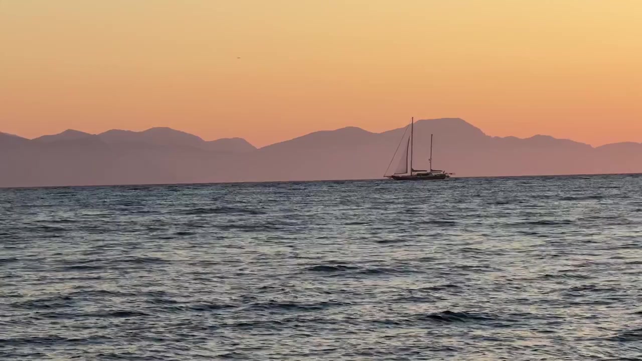 Filmisk vidvinkelbillede af en sejlbåd, der glider hen over roligt vand ved solnedgang, med varmt gyldent lys og bjergsilhuetter, der skaber en fredfyldt naturskøn baggrund.