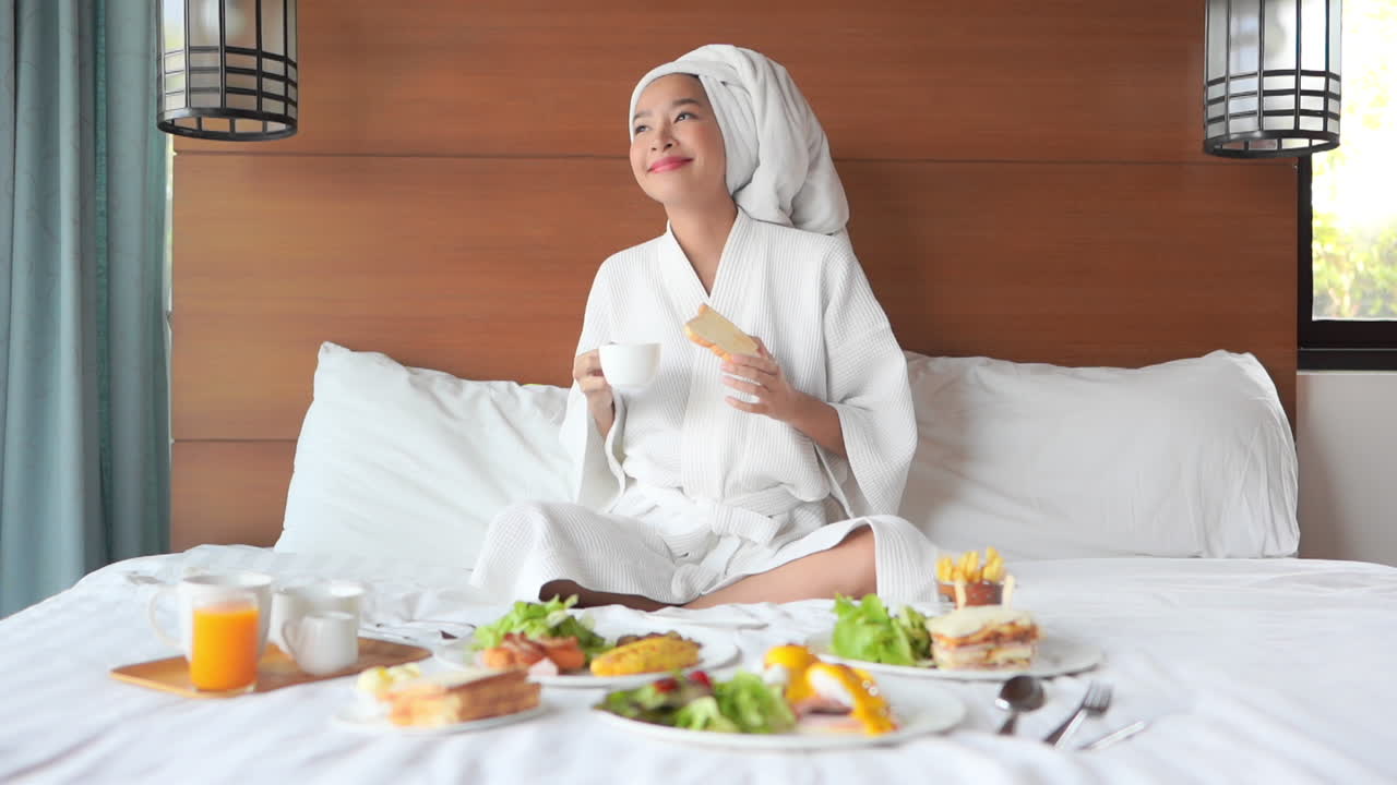 A woman fresh from a bath and wrapped in a robe, surrounded by a banquet of brunch food spread out across her hotel bed, sips coffee