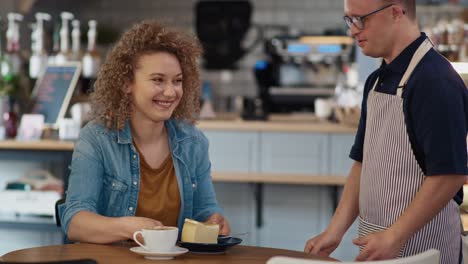 Caucasian-male-waiter-with-down-syndrome-serving-a-piece-of-cake-in-the-cafe-to-the-client-at-the-table.