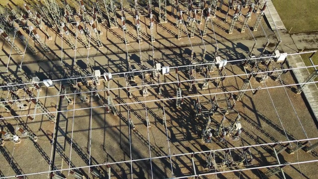 Drone footage slowly panning over a large electrical substation with complex networks of wires, transformers, and switchgear casting long shadows on the ground during a sunny day