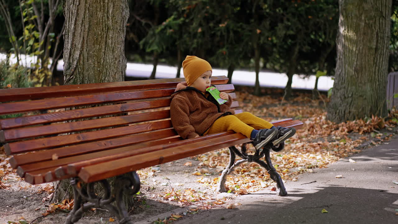 Approaching the little kid sitting on the bench in the park. Cute baby having a snack on the walk.