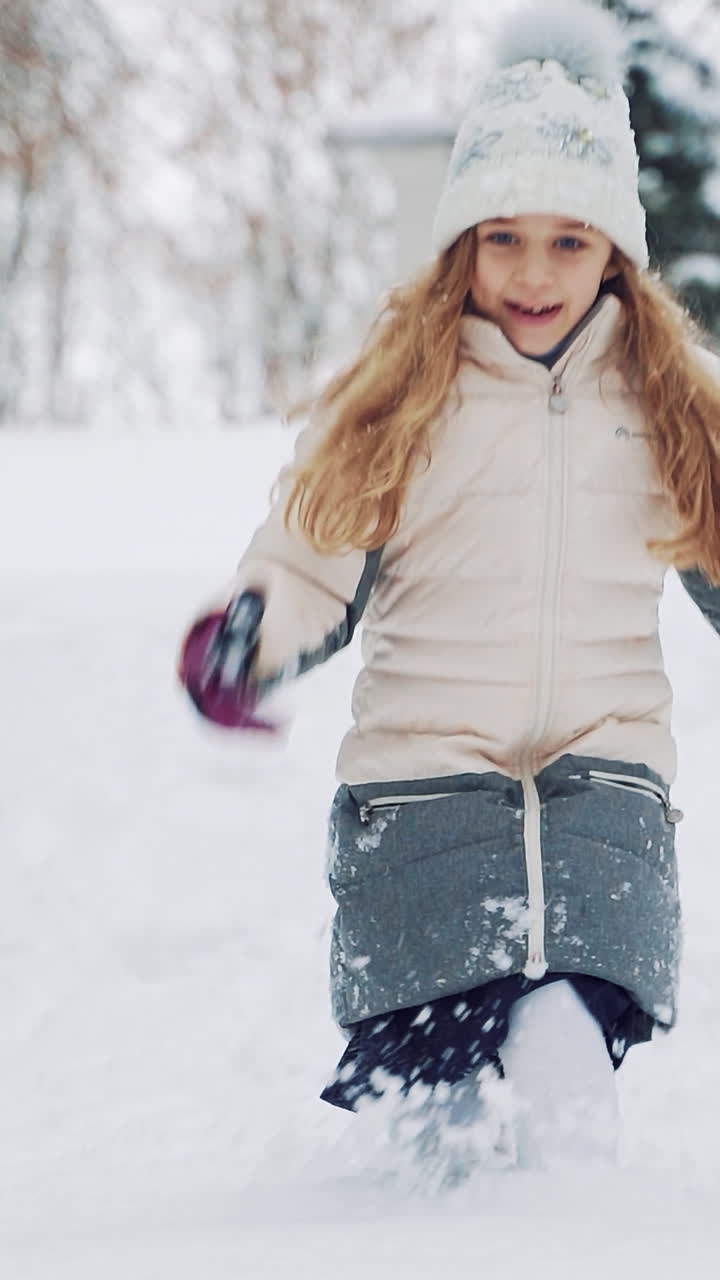 A little girl in warm clothes is descending from the hill and touching the snow. The child is scattering snow by her hands on the background of snowfall in the park. Slow motion Vertical video