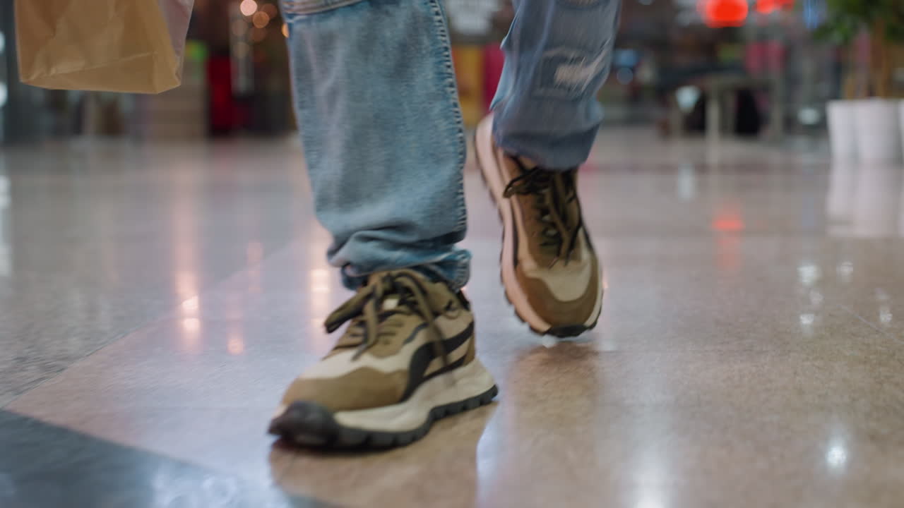 Close up of person legs in blue ripped jeans and sneakers walking across shiny indoor floor while carrying shopping bags, creating dynamic motion and lifestyle atmosphere with blurred background