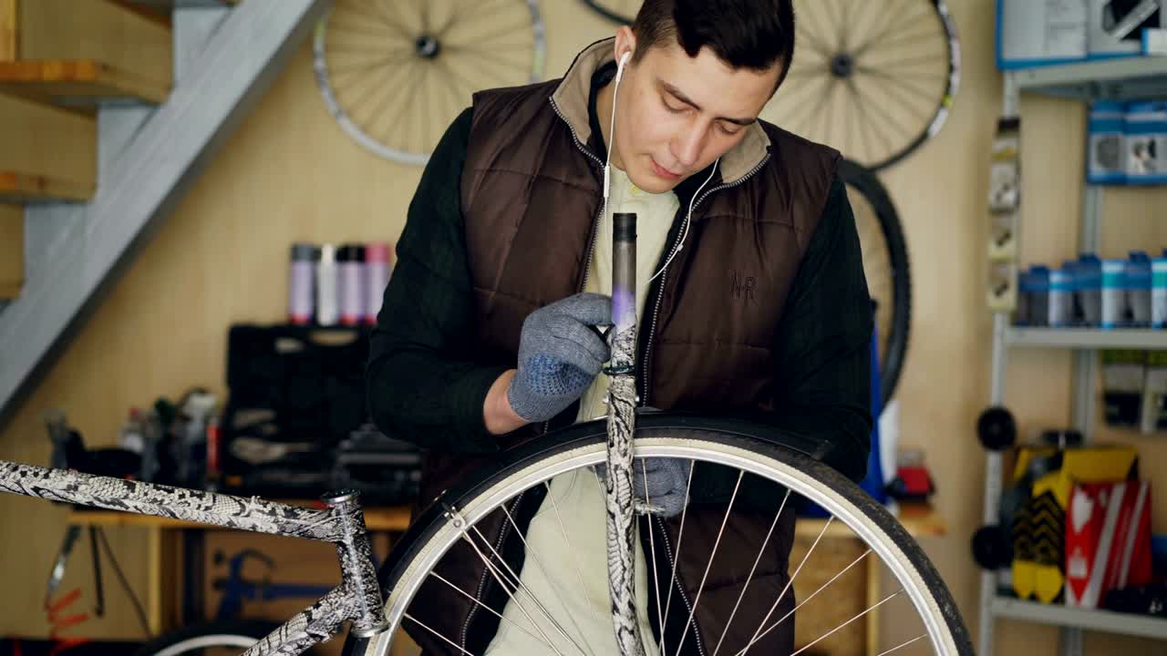 el joven mecánico concentrado está engrasando la rueda de la bicicleta y escuchando música con auriculares mientras repara la bicicleta en su pequeño estudio casero.