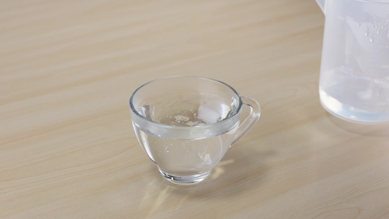Clear liquid pours from a plastic pitcher into a glass mug on a wooden table.