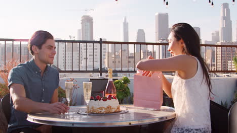 man giving woman gift and card as they celebrate on rooftop terrace with city skyline in background