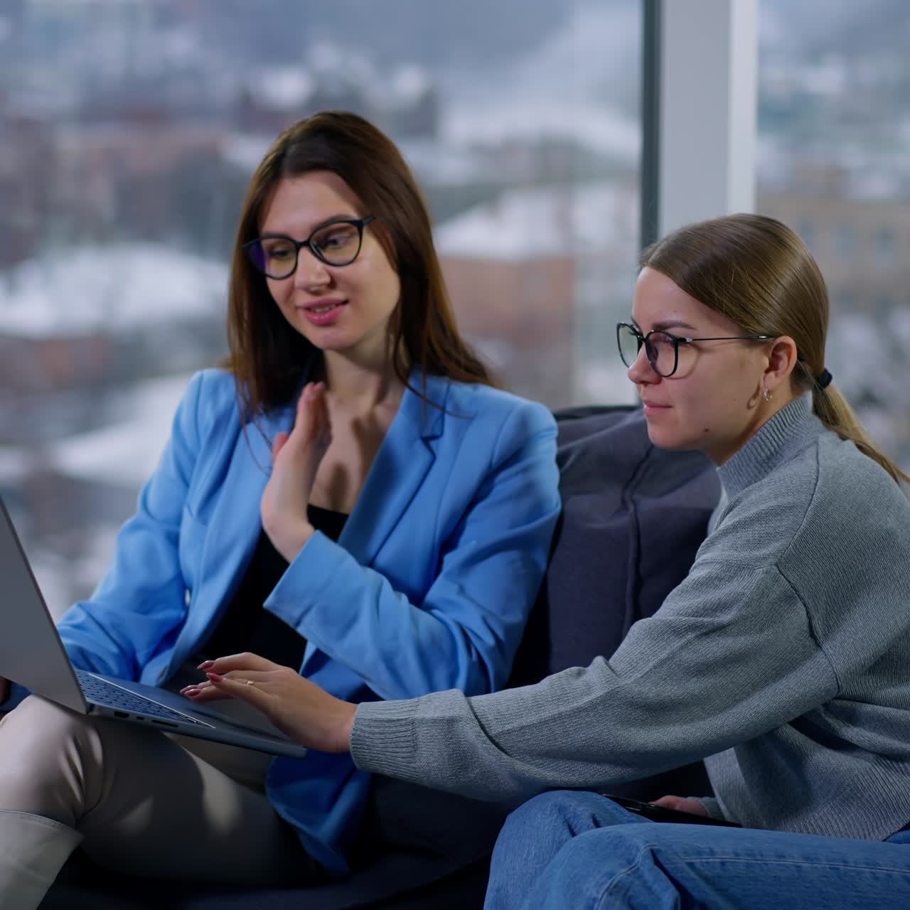 Communicating female colleagues sit in soft armchairs discussing files on laptop. Co-working in the office