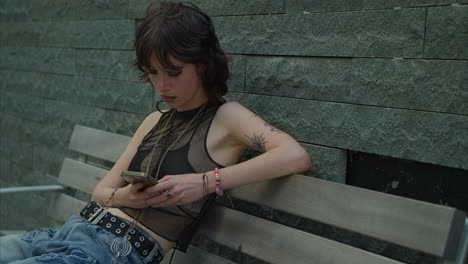 retrato de moda al aire libre de una mujer joven de estilo alternativo con tatuajes sentada en un banco usando un teléfono móvil