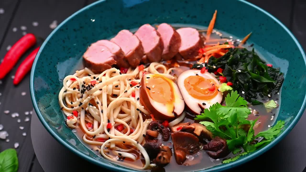 青いボウルに入った、卵と野菜のおいしい麺スープ。ボウルには麺、スライスされた肉、野菜とハーブを添えた半熟卵が入っています。