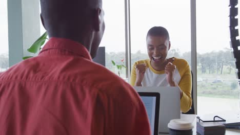 Happy-african-american-creative-businesswoman-celebrating-at-desk-in-office-with-male-colleague