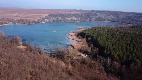 Aerial-landscape-forward-shot-of-barely-frozen-lake-with-reeds-and-pinetrees,-Szűcsi,-Hungary,-Europe