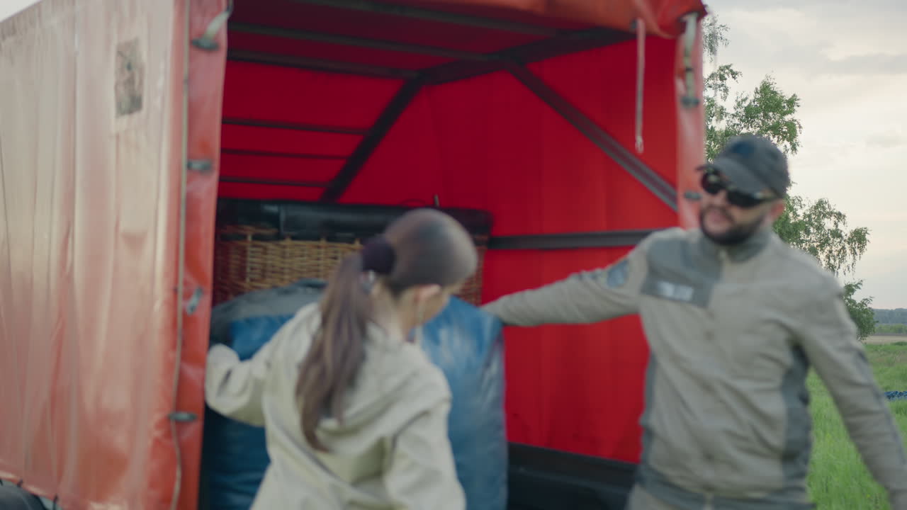 Team of friends unloads large blue gear from open truck trailer with wicker basket visible, preparing equipment for ballooning activity near wide field under soft evening sky, teamwork in motion