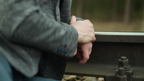 eine nahaufnahme von händen eines mannes, die zusammengeklemmt auf einer eisenbahnstrecke ruhen. er trägt einen grauen blazer und blaue jeans