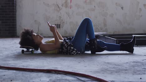 mixed race woman in an empty building lying on a skateboard using smartphone laughing