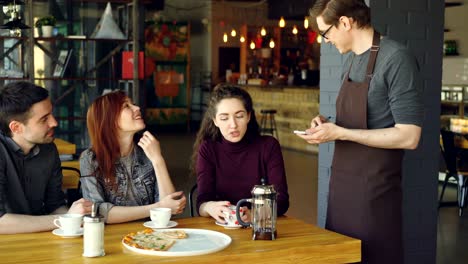 friendly male waiter is taking order from cheerful group of friends sitting at table in cafe and talking. eating out, food and drink industry and friendship concept.