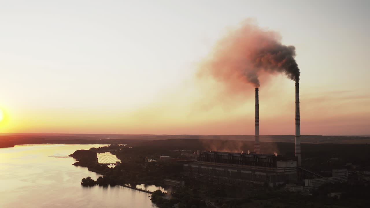 Harmful factory near the river at dusk. Industrial zone with chimneys pouring dark smoke into the air at sunset. Pollution of the environment. Aerial view.