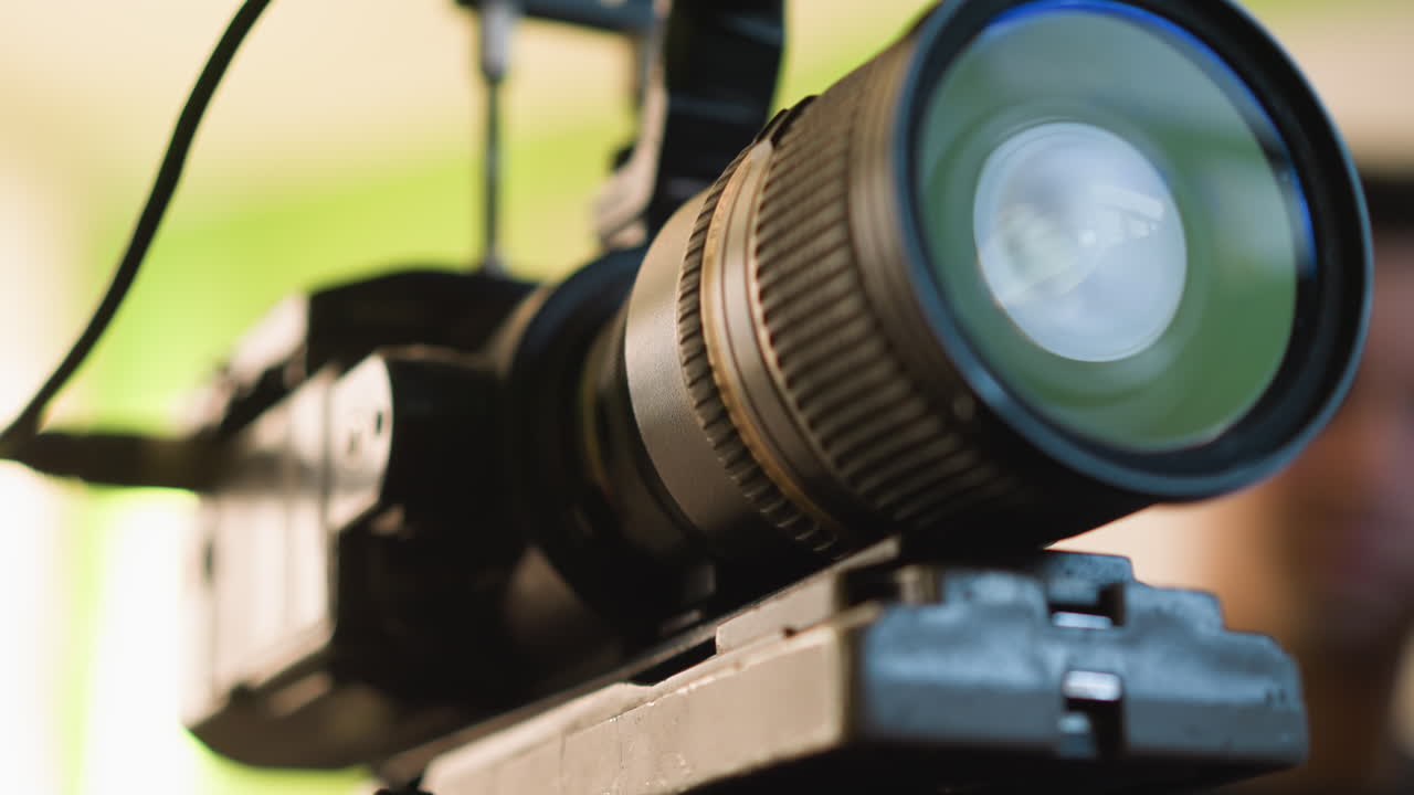 Close-up of camera tripod quick release plate with blurry camera body in background. Green backdrop with soft indoor lighting and focus on plate. Hand is near plate adjusting position