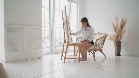 woman artist draws picture using easel sitting in armchair