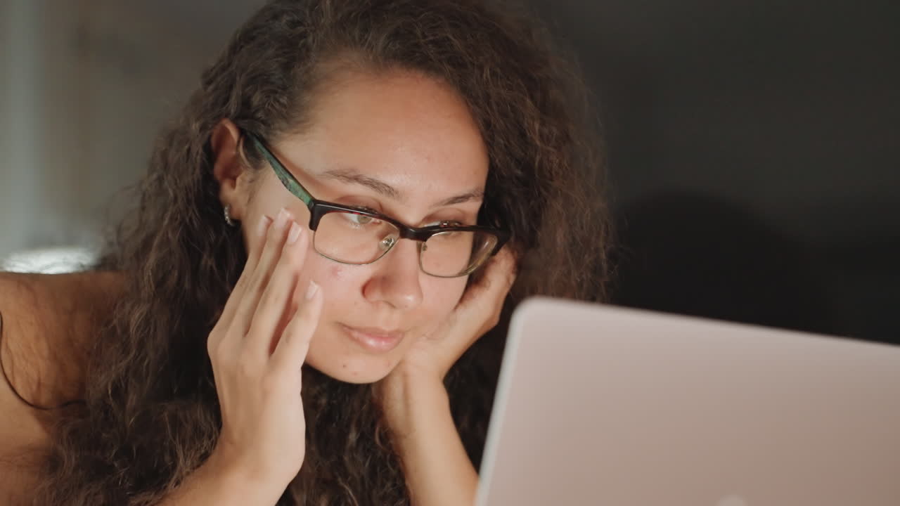 Fatica da laptop a tarda notte e quieta riflessione in camera d'hotel, gesti stanchi e mano sul viso, bagliore fioco della lampada su occhiali e capelli ricci, umore stanco di un professionista solitario o di un viaggiatore che bilancia il lavoro.
