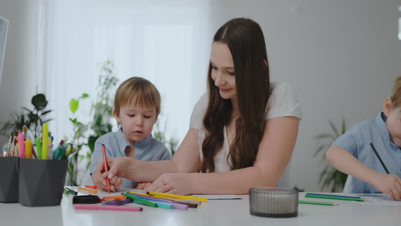 una madre joven con dos hijos sentados en una mesa blanca dibuja lápices de color en papel ayudando a hacer la tarea