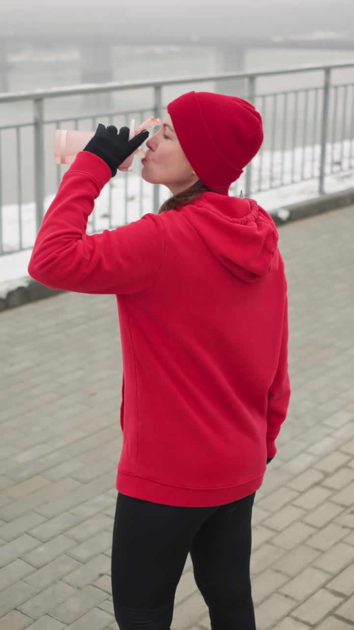lady in red hoodie and beanie sipping water from pink bottle outdoors during serene winter day with snowy ground, iron railing, calm river, and distant misty bridge in peaceful, tranquil setting