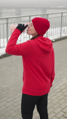lady in red hoodie and beanie sipping water from pink bottle outdoors during serene winter day with snowy ground, iron railing, calm river, and distant misty bridge in peaceful, tranquil setting