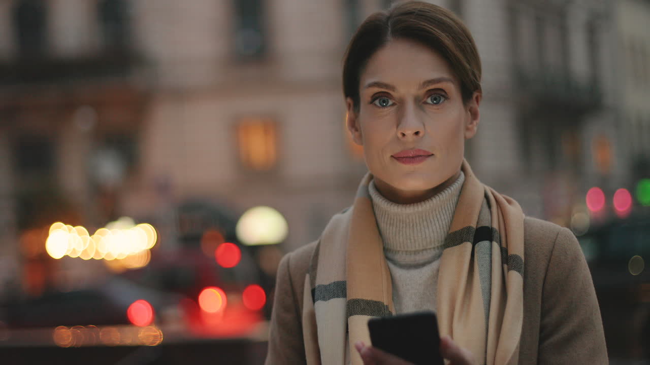 крупный план молодой кавказской деловой женщины, пишущей смс на смартфоне на улице с городскими огнями на заднем плане, затем она смотрит в камеру и улыбается