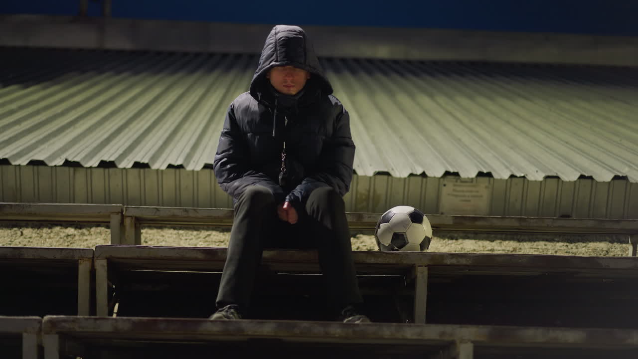검은 옷과 후드 재을 입은 남자가 빈 경기장에서 혼자 앉아 있으며, 에 축구공이 놓여 있습니다.