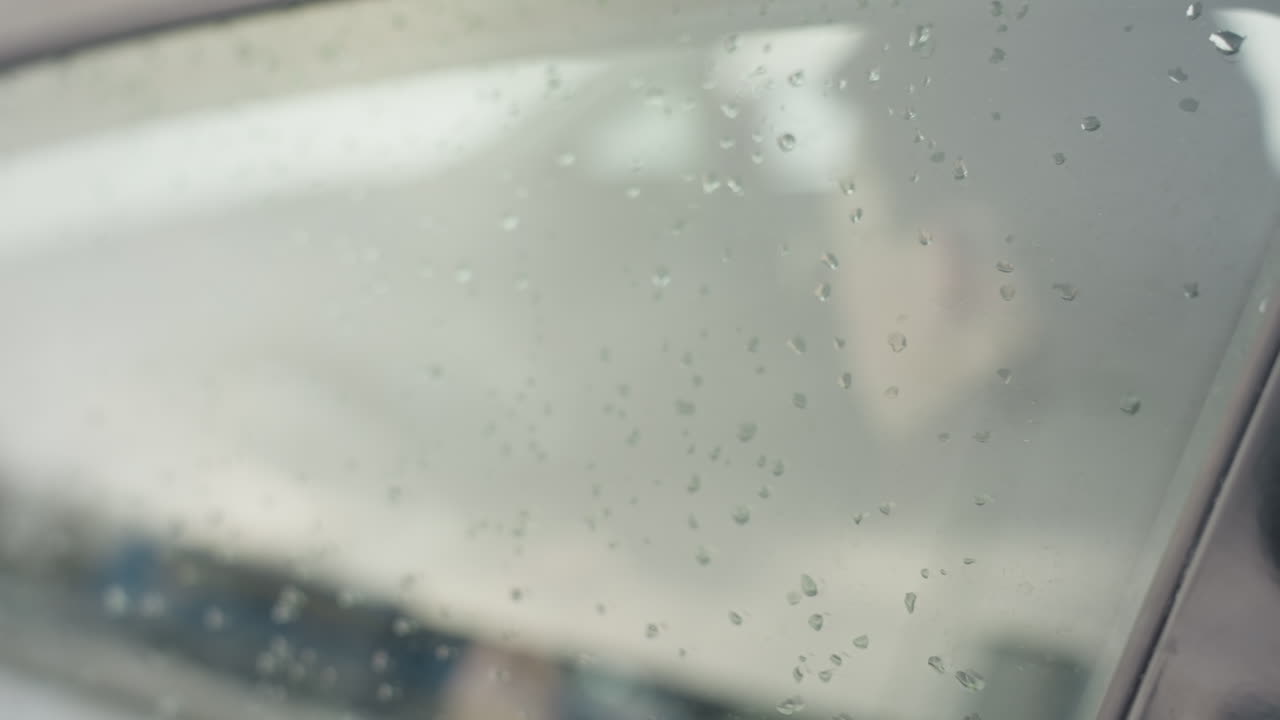 close up of car window covered in water droplets on rainy day with blurred view of person inside vehicle performing activity, creating moody urban atmosphere and soft exterior reflections