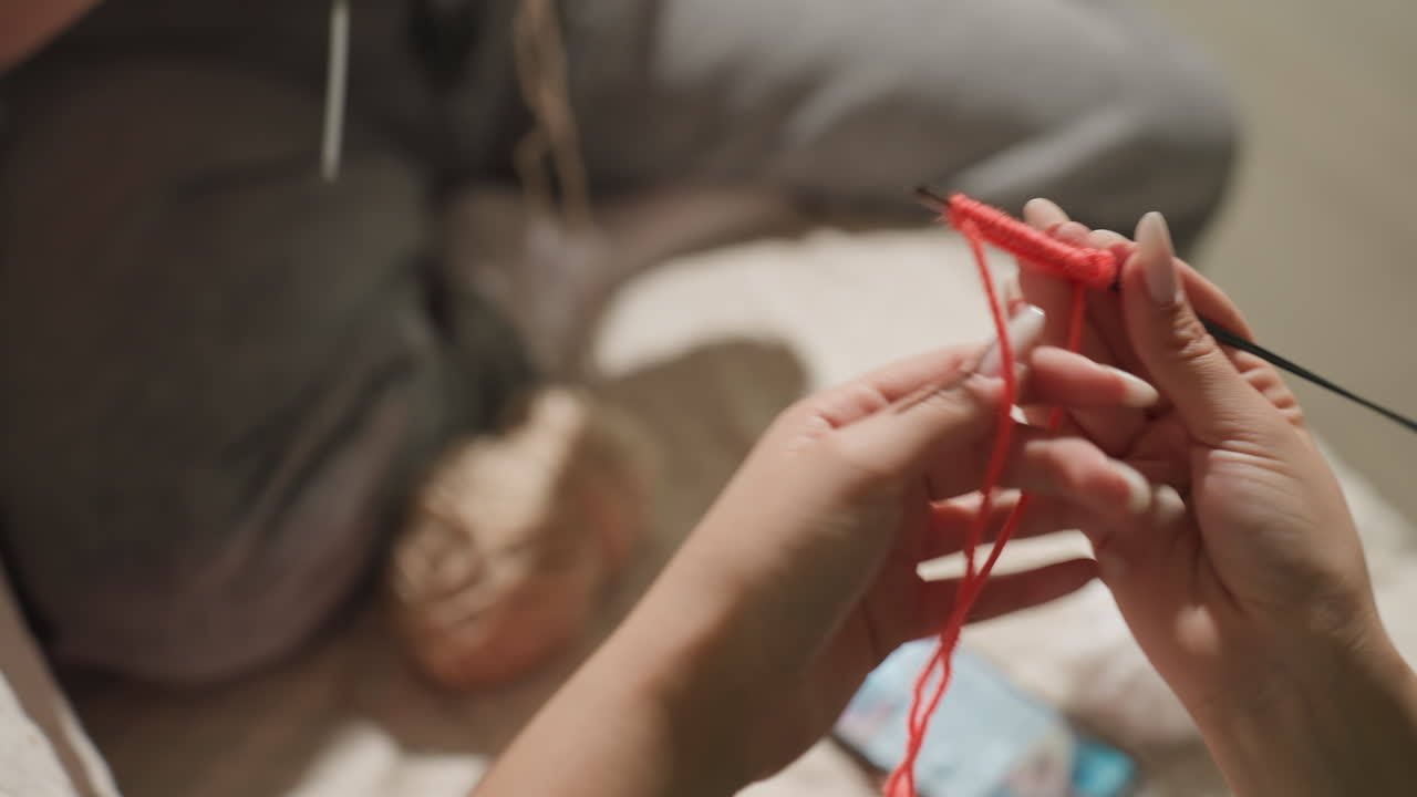 저녁에 따뜻한 조명 아래에서 공예 시간을 보내는 모습이에요. 은은하고 따뜻한 빛 아래에서 한 여성이 조용히 뜨개질을 하고 있어요. 붉은색 털실과 바늘로 부드러운 저녁 조명 아래에서 차분하게 뜨개질을 하는 여성의 일상이 돋보이네요.