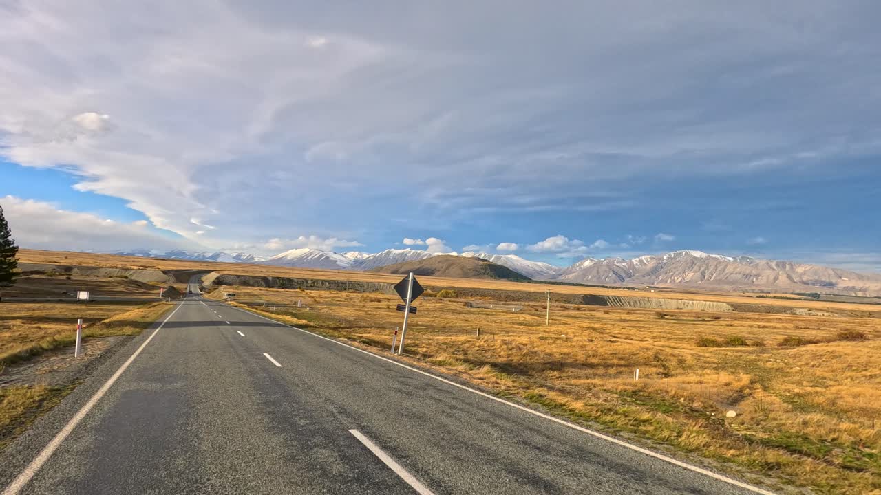 Forward-facing dashcam view of a car driving on an empty highway through golden autumn fields, with distant mountains and dramatic sky, in soft daylight