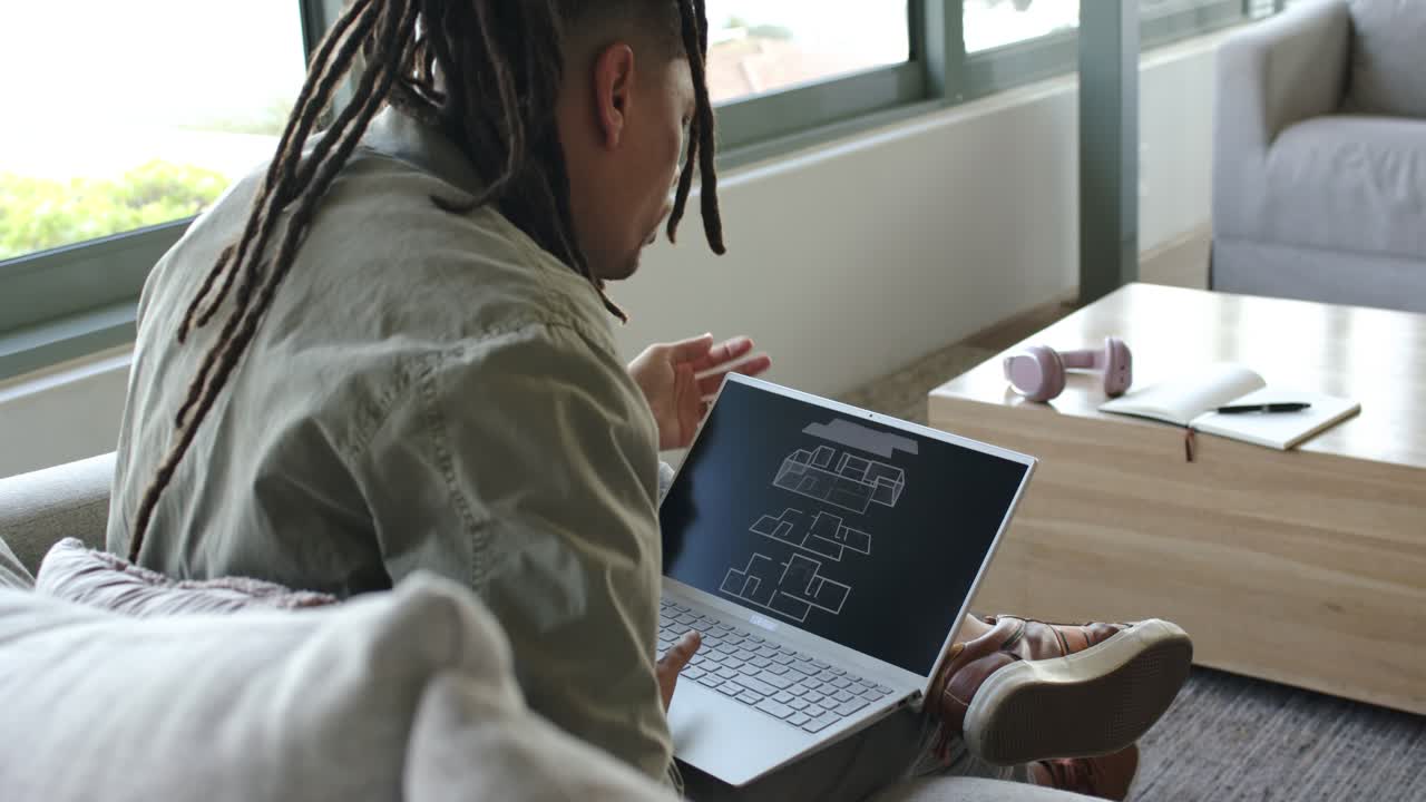 Man is lifting laptop onto lap and opening it, pushing dreadlocks back, examining blueprint on sofa