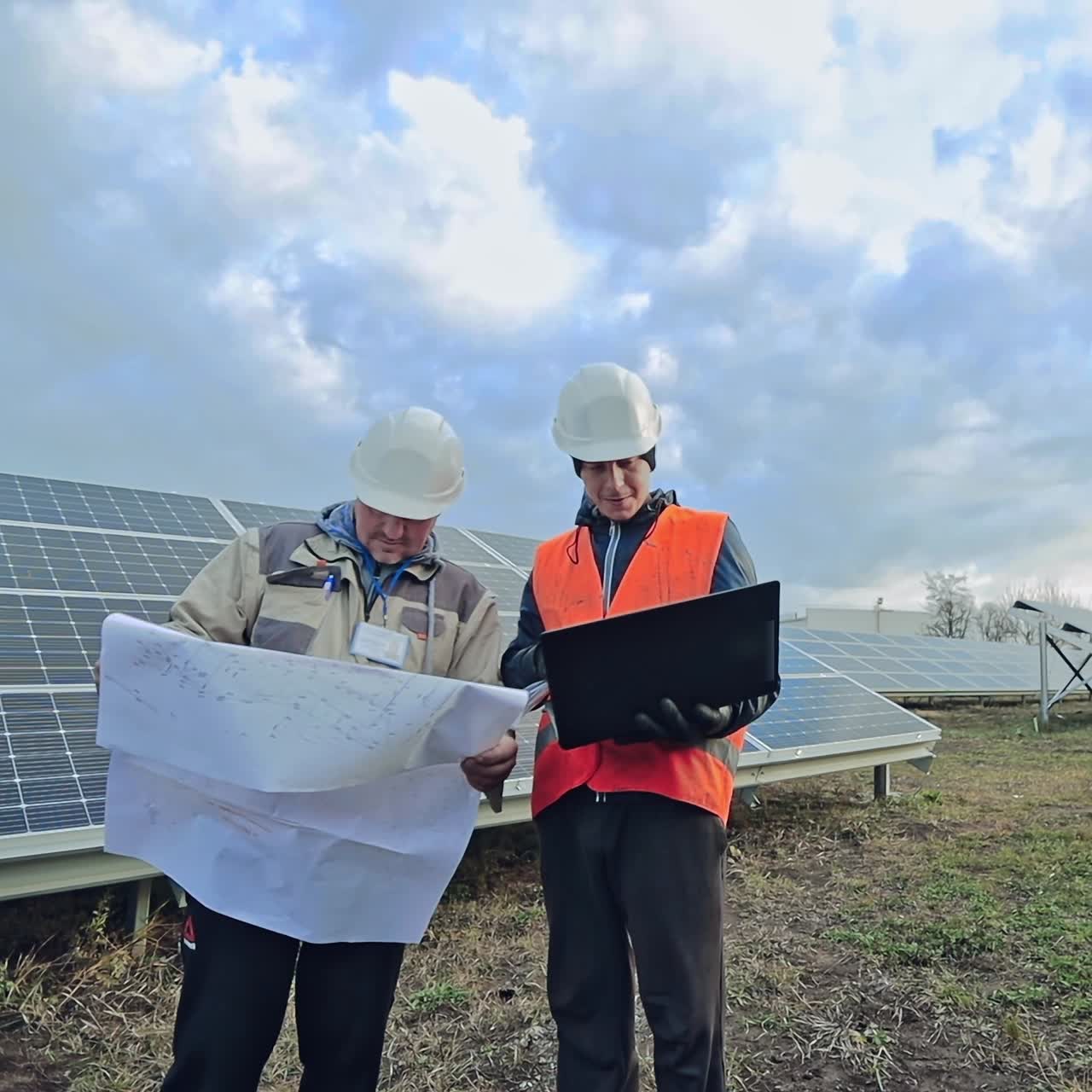 Technicians look into laptop and project plan on the solar farm. Two workers in protective helmets on the background of solar panels.