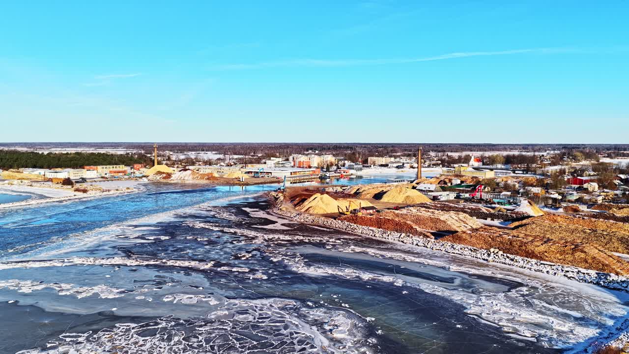 Hafen Salacgrīva in Lettland mit gefrorener Flussmündung und Materiallagern, Holzstämme im Industriehafen in Luftaufnahme an einem sonnigen Wintertag