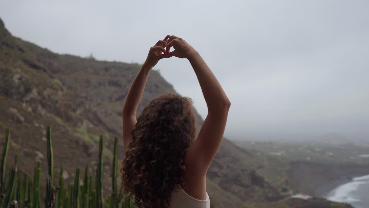 A woman sits on a mountain's crest, meditating and expressing the Maha Sakal hand gesture, while the ocean and green mountains envelop her