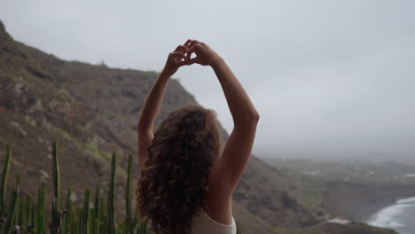 una mujer se sienta en la cresta de una montaña, meditando y expresando el gesto de la mano maha sakal, mientras el océano y las montañas verdes la envuelven