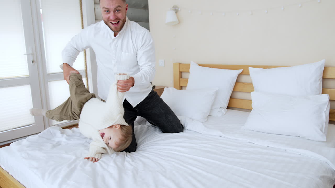 Happy Caucasian male takes kid's hand and foot waving him from side to side. Father tosses his son on the pillows. Woman at backdrop.