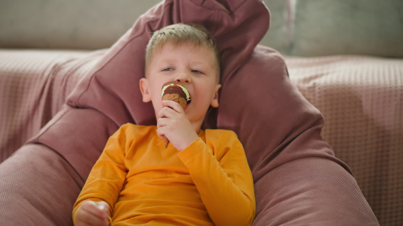Little boy relaxing on cozy pink bean bag chair with eyes closed while enjoying chocolate-covered ice cream cone, wearing bright yellow shirt and cream pants, surrounded by soft homey background