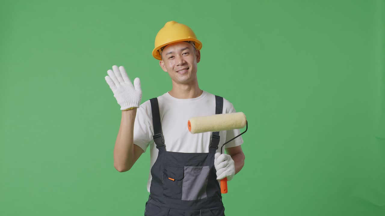 Asian Man Painter Wearing Safety Helmet Smiling, Waving Hand, And Saying Bye While Standing In The Green Screen Background Studio