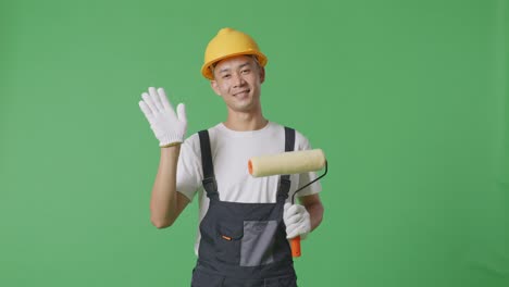 Asian Man Painter Wearing Safety Helmet Smiling, Waving Hand, And Saying Bye While Standing In The Green Screen Background Studio