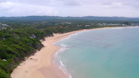 Vuele-Sobre-El-Agua-Tranquila-Del-Océano-En-La-Orilla-Arenosa-En-El-Paisaje-Natural-En-Byron-Bay,-Costa-De-Australia