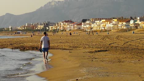 Man-at-beach-walking-along-shoreline-of-coastal-town,-slow-motion-wide-shot