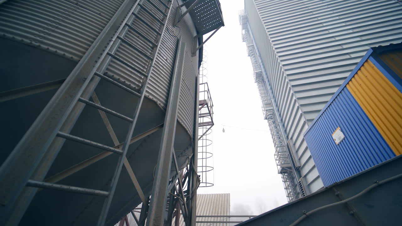 Huge silo tank with the metal ladder and a little balcony. A turning view from the grain elevator tank to other premises and cisterns in the agricultural plant.