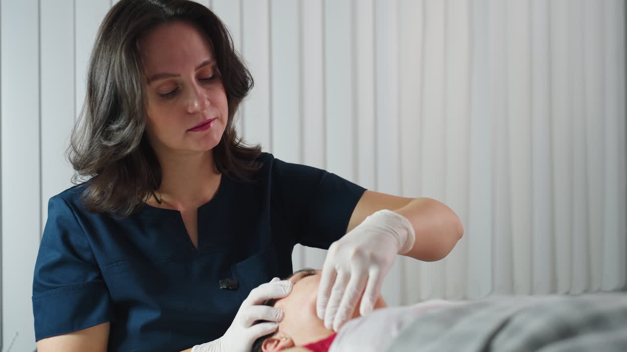 Wellness technician wearing disposable gloves dips fingernail inside mouth of woman while other hand cradles jawline smoothly during session in spa clinic gentle professional care and relaxation