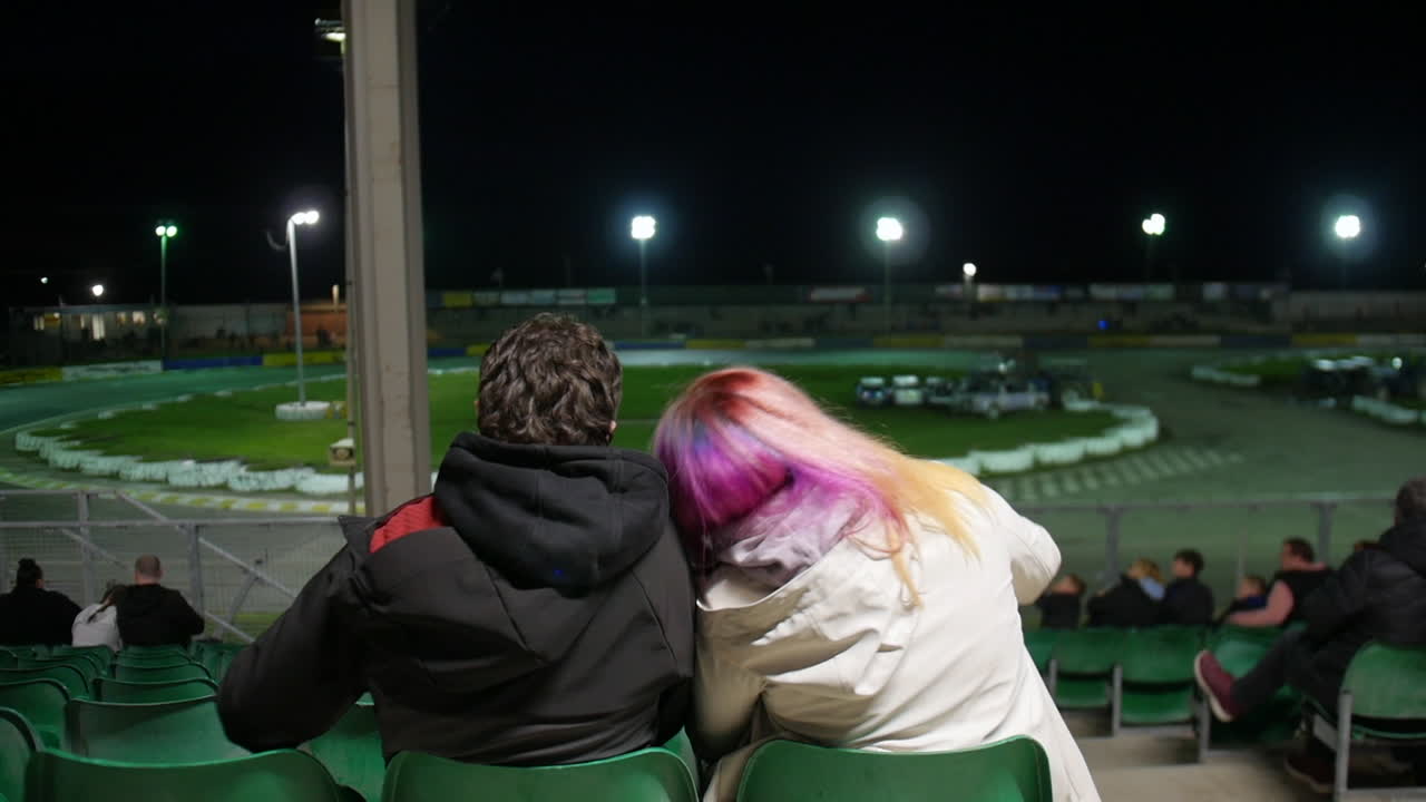 A couple sitting closely in green stadium seats, with one resting their head on the other's shoulder, watching the illuminated racetrack at night. A quiet, intimate moment