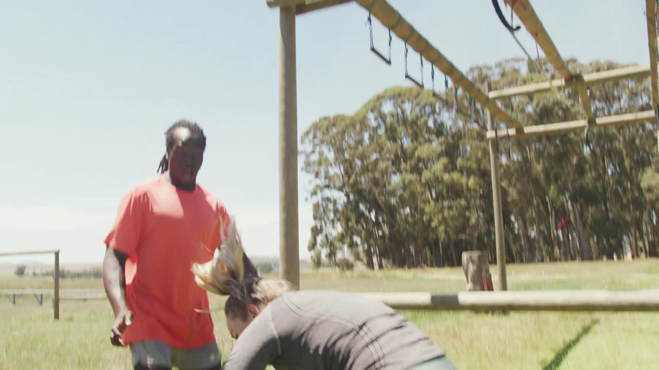 mujer caucásica en forma cayendo de las barras en una carrera de obstáculos ayudada por un colega afroamericano